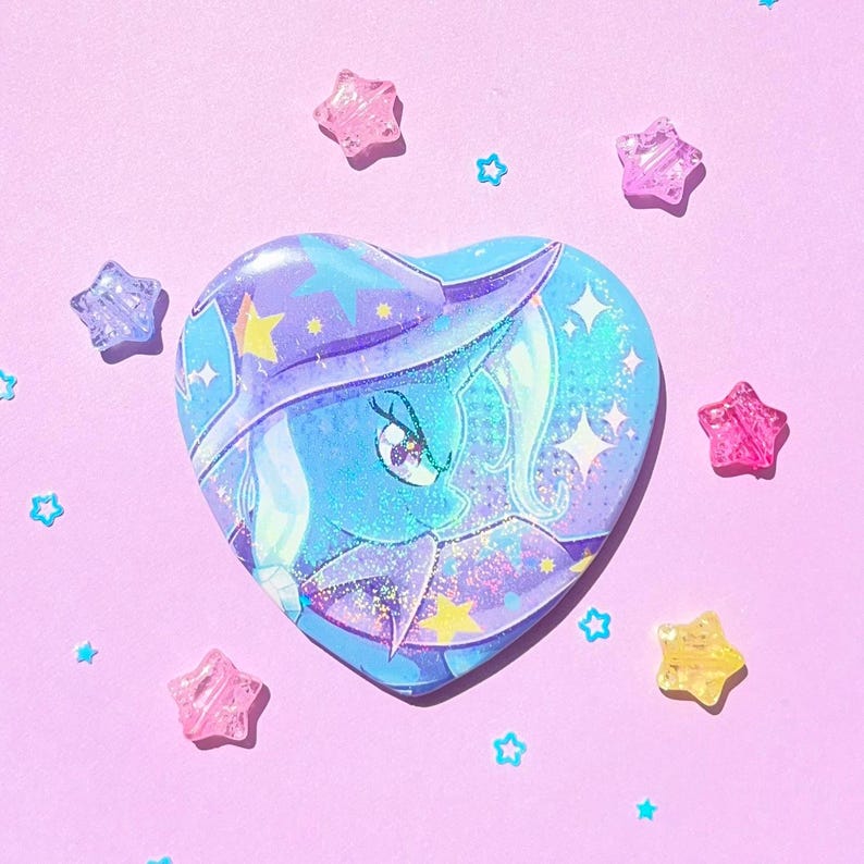 May include: A heart-shaped button featuring a blue and purple illustration of a pony with a star-studded design. The button is surrounded by star-shaped and star-shaped confetti on a pink background.