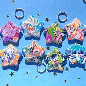 May include: A collection of star-shaped character pins featuring various characters from the Sonic the Hedgehog franchise. The pins are colorful, with detailed illustrations and a blue background. Gold rings and star-shaped confetti are scattered around the pins.