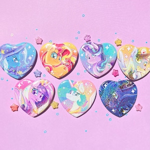 May include: A collection of heart-shaped buttons featuring colorful illustrations of cartoon characters. The buttons have a glossy finish and are decorated with stars and glitter. The background is a light purple color.