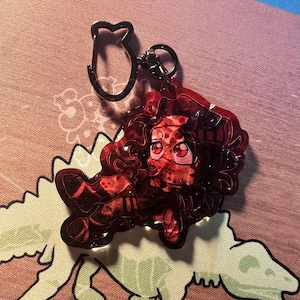 Op de afbeelding: Een doorschijnende rode acryl sleutelhanger met een gestileerd stripfiguur met donker haar en grote ogen. De bedel is bevestigd aan een zwarte metalen sluiting in de vorm van een kat. Het ontwerp heeft een glanzende afwerking met glitter.