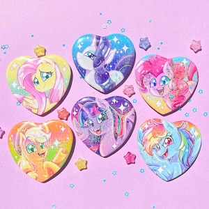 May include: Six heart-shaped pins featuring colorful illustrations of My Little Pony characters. Each pin has a different pony design with glitter accents. The pins are set against a light pink background with star and star-shaped confetti.