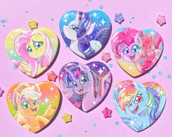 May include: Six heart-shaped badges featuring My Little Pony characters. Each badge has a different pony design with glitter accents. The badges are set against a light pink background with star and star-shaped confetti.