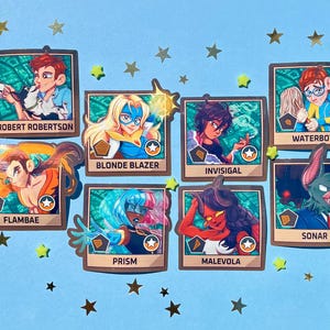 Puede incluir: Una colección de siete tarjetas de personajes con ilustraciones de varios superhéroes y villanos. Cada tarjeta presenta un diseño de personaje único, un nombre y un emblema de estrella. Las tarjetas están dispuestas sobre una superficie azul claro con confeti dorado en forma de estrella.