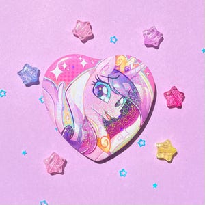 May include: A heart-shaped button featuring a cartoon unicorn with a crown, in shades of pink, purple, and white. The button is surrounded by star-shaped and star-shaped confetti on a light purple background.