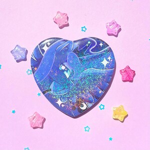 May include: A heart-shaped sticker featuring a blue illustration of a winged creature with a starry background. The sticker is surrounded by pink, purple, and yellow star-shaped embellishments on a pink background.