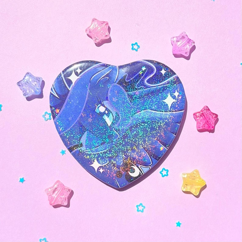 May include: A heart-shaped sticker featuring a blue illustration of a winged creature with a starry background. The sticker is surrounded by pink, purple, and yellow star-shaped embellishments on a pink background.