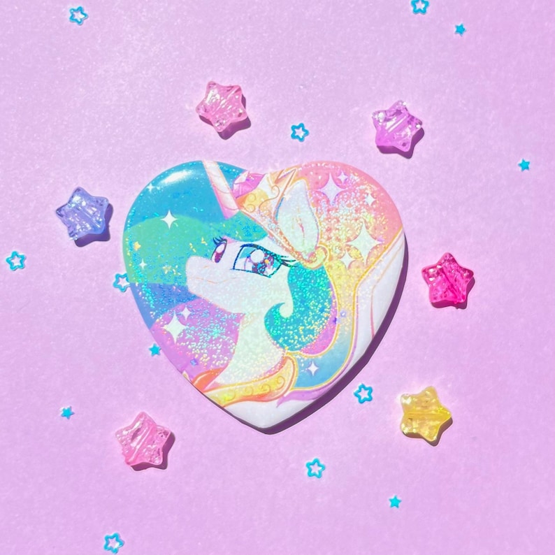 May include: A heart-shaped button featuring a colorful illustration of a unicorn with a crown and flowing mane. The button has a holographic effect with blue, pink, and yellow hues. Scattered around the button are star-shaped and star-shaped beads on a lavender background.
