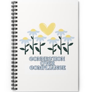 以下が含まれることがあります： 黄色のハートと青い花が描かれた白いスパイラルノート。"CONNECTION OVER COMPLIANCE"というテキストが青い文字で印刷されています。