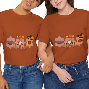 Puede incluir: Dos personas con camisetas marrones con un diseño de temática de Acción de Gracias que incluye un gnomo, un balón de fútbol y girasoles. El diseño incluye el texto "ABA".