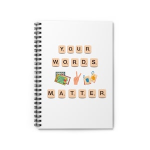 以下が含まれることがあります： 白いノートにスパイラルバインディングが施され、ボードゲームの文字タイルを使って「YOUR WORDS MATTER」というフレーズが綴られています。タイルは白文字と数字が書かれた茶色です。フレーズはノートの中央に配置されています。ゲームボード、ピースサインをする手、2つのサムズアップアイコンの画像もあります。