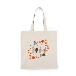 Puede incluir: Una bolsa de tela beige con un diseño de hojas de otoño y el texto "its fall yall" en letras negras. La palabra "fall" está representada en el lenguaje de señas americano.