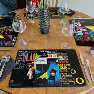 May include: A table setting with four place settings, each with a black napkin, silverware, and a wine glass. The placemats are made from vintage vinyl record covers with colorful designs and text. The center of the table has a vase with red and white flowers.