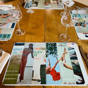 May include: A table setting with two wine glasses, two place settings, and a placemat with a collage of vintage record covers featuring images of women and the text "Songs Ruby & Peg Anna & Tam Thompson's Gin' and "Kapp Records"
