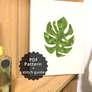 Pode incluir: Uma obra de arte emoldurada com um design geométrico de folha de monstera verde sobre um fundo branco. A obra de arte é exibida em uma prateleira de madeira. O texto na imagem diz "PDF Pattern + stitch guide".