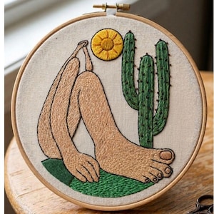 May include: A circular wooden embroidery hoop containing a fabric panel with a hand-stitched design. The embroidery depicts a tan-colored figure sitting with knees drawn up, a bright yellow sun, a green cactus, and a patch of green ground.
