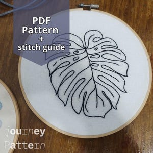 Pode incluir: Bastidor de bordado com um desenho de folha de Monstera costurada em preto sobre tecido branco. O texto "PDF Pattern + stitch guide" é visível. O bastidor de madeira está sobre uma superfície de madeira. As palavras "journey" e "Pattern" também são visíveis.