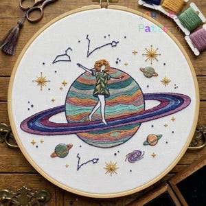 Pode incluir: Um bastidor de bordado com fundo de tecido branco apresentando um design costurado à mão de uma pessoa em pé sobre um planeta grande e multicolorido com anéis. A cena inclui planetas menores, estrelas e constelações bordadas.