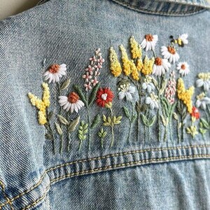 以下が含まれることがあります： 刺繍の花柄が施されたライトブルーのデニムジャケット。刺繍には白いデイジー、黄色のワイルドフラワー、赤い花、小さな蜂が描かれています。ジャケットの背面には、カラフルなステッチの花が飾られています。
