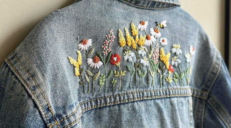 以下が含まれることがあります： 刺繍の花柄が施されたライトブルーのデニムジャケット。刺繍には白いデイジー、黄色のワイルドフラワー、赤い花、小さな蜂が描かれています。ジャケットの背面には、カラフルなステッチの花が飾られています。