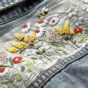 以下が含まれることがあります： デニムジャケットのディテール。白いデイジー、黄色い花、赤い花、緑の葉が刺繍されています。小さな蜂も刺繍されています。ジャケットは水色です。