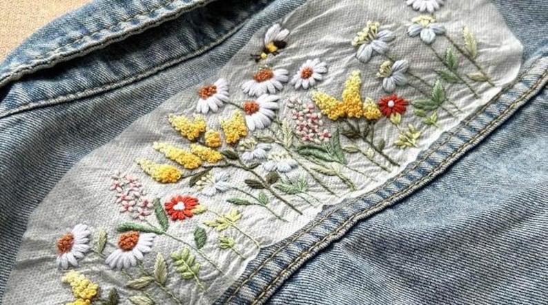 以下が含まれることがあります： デニムジャケットのディテール。白いデイジー、黄色い花、赤い花、緑の葉が刺繍されています。小さな蜂も刺繍されています。ジャケットは水色です。