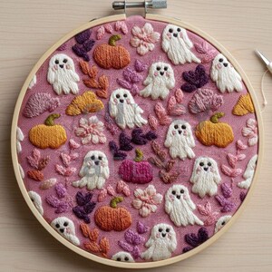 May include: Hand-stitched embroidery art in a wooden hoop. The design features white ghosts, orange pumpkins, and pink flowers on a pink fabric background. The embroidery is detailed with various colors of thread, creating a textured and whimsical Halloween theme.