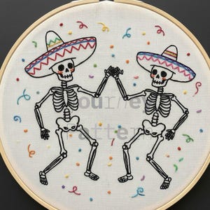 May include: An embroidered hoop art piece featuring two dancing skeletons wearing sombreros. The skeletons are black and white, holding hands, and surrounded by colorful confetti. The embroidery is on a white fabric, set in a wooden hoop.