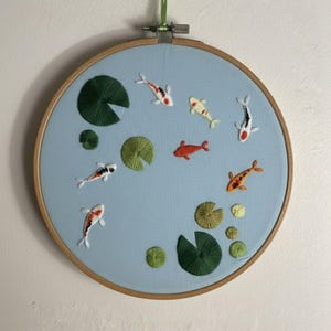Könnte beinhalten: Gesticktes Kunstwerk mit bunten Koi-Fischen und grünen Seerosen auf hellblauem Hintergrund. Die Stickerei ist in einem runden Holzreifen angebracht und eignet sich als dekoratives Wandbild.