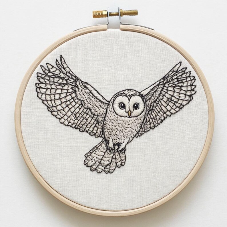 May include: Embroidery hoop art featuring a detailed black and white owl with outstretched wings. The owl is centered on a white fabric, set within a natural wood hoop. The artwork showcases intricate stitching, capturing the owl's feathers and facial features.