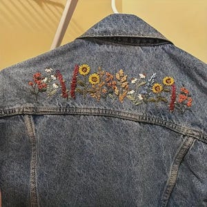 以下が含まれることがあります： 背中上部に花の刺繍が施されたデニムジャケット。刺繍は、ヒマワリ、デイジー、その他のワイルドフラワーを黄色、オレンジ、赤、白の色合いで表現しています。ジャケットはミディアムウォッシュブルーです。