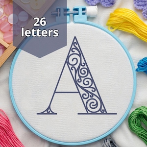 May include: An embroidery hoop with a light blue frame holds a white fabric with a dark blue letter "A" design. The design is surrounded by colorful embroidery floss in pink, yellow, blue, green, and purple.