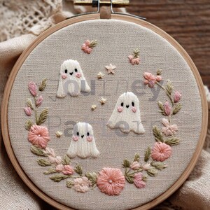 May include: A handmade embroidery hoop featuring three white ghost figures with blush cheeks. The ghosts are surrounded by a wreath of pink and green embroidered flowers and leaves. The background is a light beige fabric.
