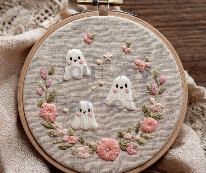 May include: A handmade embroidery hoop featuring three white ghost figures with blush cheeks. The ghosts are surrounded by a wreath of pink and green embroidered flowers and leaves. The background is a light beige fabric.