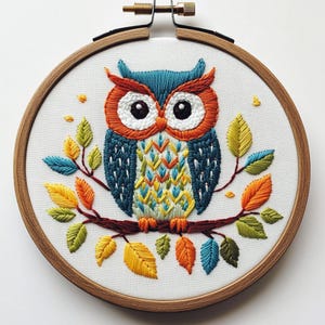 May include: A colorful owl embroidery on white fabric, set in a wooden hoop. The owl features blue, orange, and white details, perched on a branch with embroidered leaves in shades of yellow, orange, and blue. A handcrafted piece.