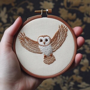 May include: Hand-stitched embroidery of a barn owl in flight, featuring brown and white thread on a cream-colored fabric. The owl is centered within a wooden embroidery hoop, showcasing detailed wing and feather patterns.