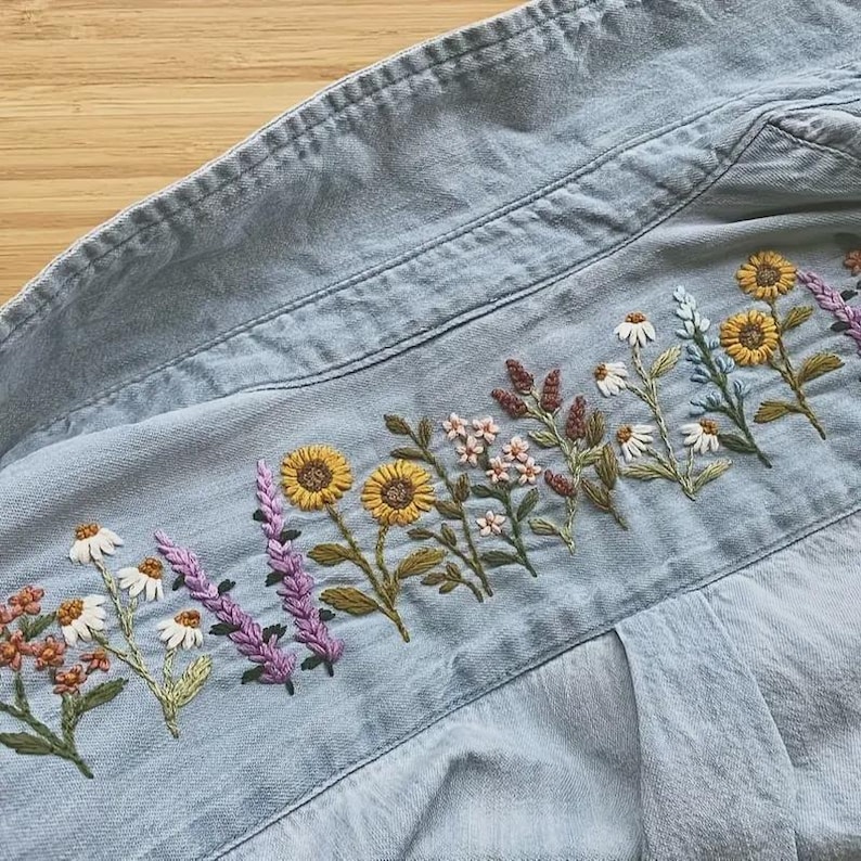 以下が含まれることがあります： 背中に詳細な花の刺繍が施された、明るいブルーのデニムシャツ。 刺繍には、ヒマワリ、デイジー、ラベンダーの小枝が、黄色、白、紫、茶色の色合いで描かれています。