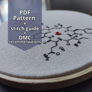 Pode incluir: Um bastidor de bordado com tecido branco com um desenho de estrutura molecular bordada em preto e um pequeno coração vermelho. O texto "PDF Pattern + stitch guide + DMC recommendations" está sobreposto na imagem.