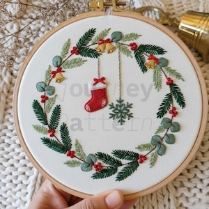 Pode incluir: Grinalda de Natal bordada num aro de madeira. A grinalda apresenta agulhas de pinheiro verdes, bagas vermelhas e pequenos sinos dourados. Uma meia vermelha e um floco de neve estão suspensos no centro. O texto "Journey Pattern" é visível.