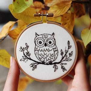May include: A hand-stitched embroidery hoop featuring a black and white owl design. The owl is perched on a branch with leaves, all set against a white fabric background. The wooden hoop is held by a person's hands, with autumn leaves in the background.