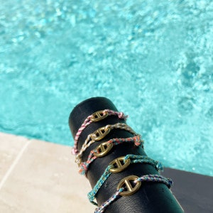 Op de afbeelding: Een stapel kleurrijke armbanden met gouden nautische bedels. De armbanden worden gepresenteerd op een zwart oppervlak.