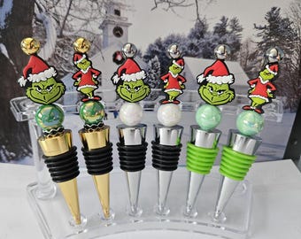May include: Six bottle stoppers featuring a green character wearing a red Santa hat. Each stopper has a decorative bead, a metal base, and black or green ribbed silicone rings. The stoppers are arranged in a clear acrylic display stand.
