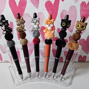 May include: A collection of six decorative pens, each topped with a different cat figurine. The pens have black, brown, and orange barrels, with silver tips. The cat figurines are in various colors and styles, including black, brown, gray, and orange.