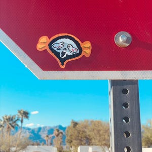May include: A sticker featuring a pixelated skull design with orange accents, affixed to a red sign. The sticker has a black outline and a white background. The sign is mounted on a metal pole with a blue sky and desert landscape in the background.