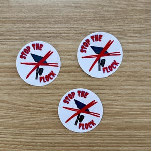 May include: Three circular white stickers featuring a graphic of a black solar-powered security camera crossed out by two red lines. The text STOP THE FLOCK is printed in red capital letters around the graphic on each of the three round stickers.