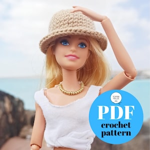 May include: A doll wearing a beige crochet hat and a white top, with a gold-colored necklace. A blue circle with white text that reads "PDF crochet pattern" is in the lower right corner. The background shows a beach scene.