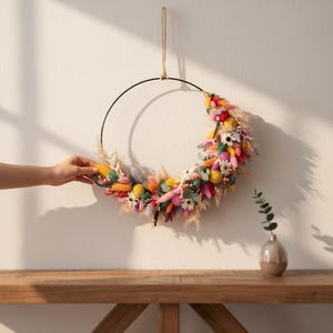 May include: A circular wall hanging featuring a black metal hoop adorned with a crescent-shaped arrangement of dried flowers and grasses in vibrant shades of pink, yellow, orange, blue, and green, suspended by a thin tan cord from the top.