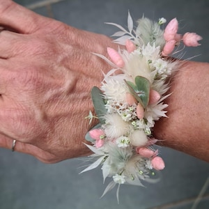 May include: A wrist corsage made with white and pink dried flowers, including fluffy white pampas grass, small white flowers, and pink blossoms. The corsage is tied with a white ribbon.