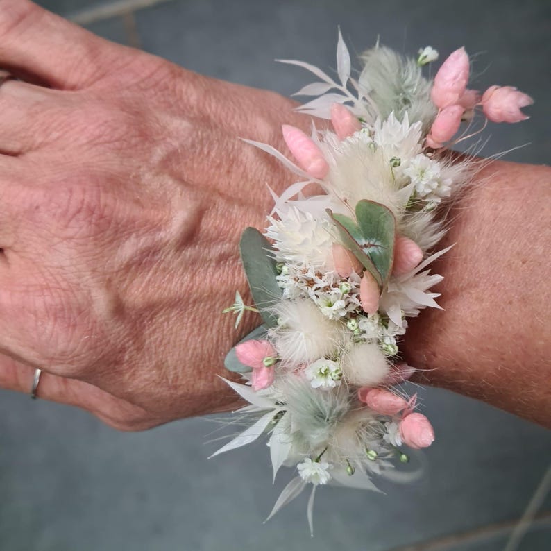 May include: A wrist corsage made with white and pink dried flowers, including fluffy white pampas grass, small white flowers, and pink blossoms. The corsage is tied with a white ribbon.