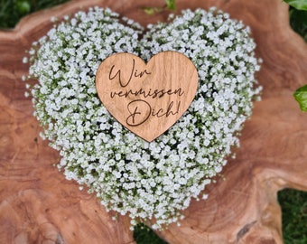 May include: A heart-shaped wooden plaque with the engraved text "Wir vermissen Dich!" resting atop a dense arrangement of small white baby's breath flowers. The wood features a visible grain pattern and a natural light brown finish.