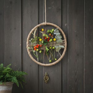 May include: A circular wall hanging featuring a thick, natural-colored rope frame. Inside the frame, thin twine is woven horizontally, supporting a collection of dried botanical elements including small red berries, yellow flowers, and green leaves.